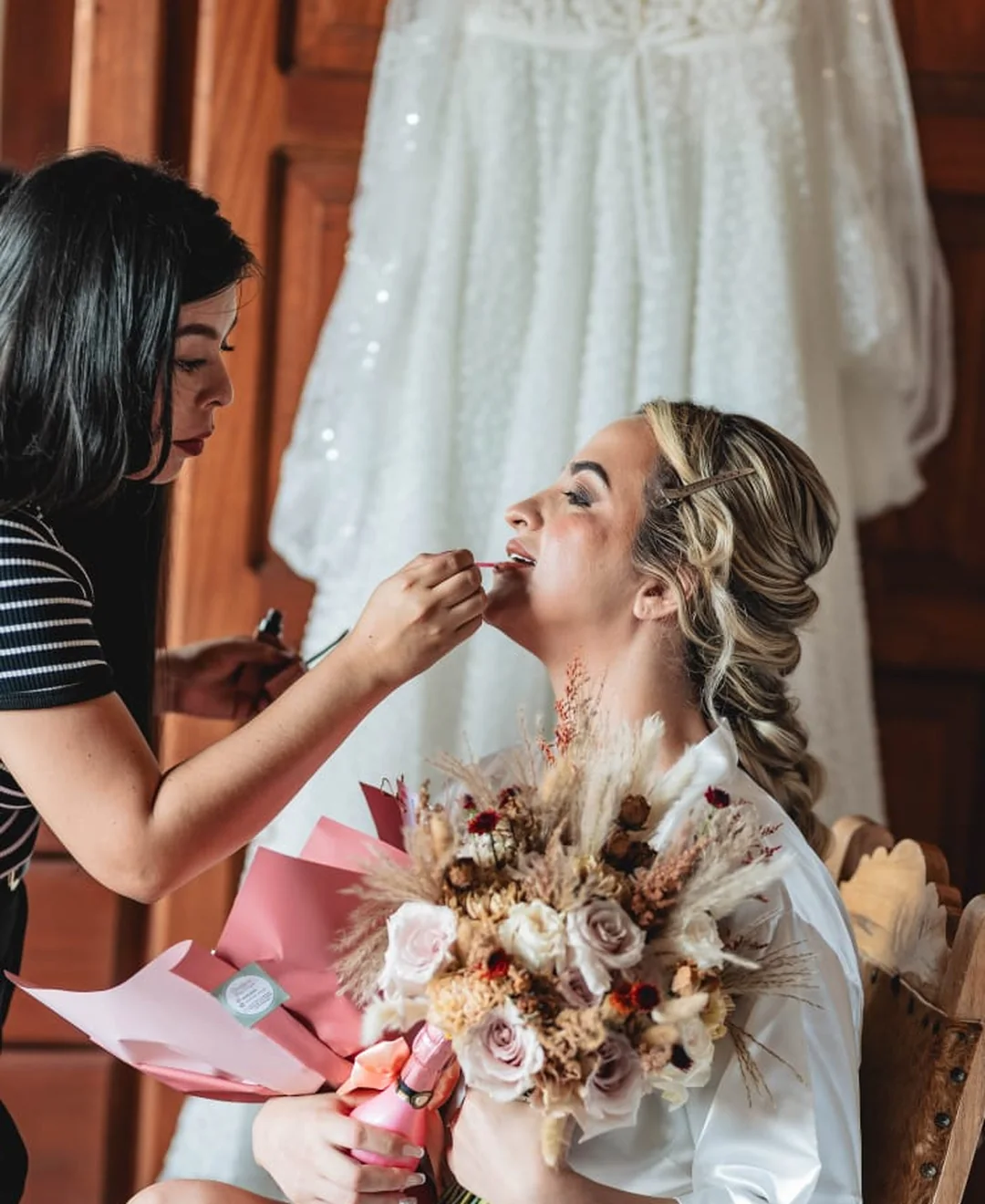 Alejandra maquillando a novia con vestido y bouquet al fondo — preparación pre-ceremonia
