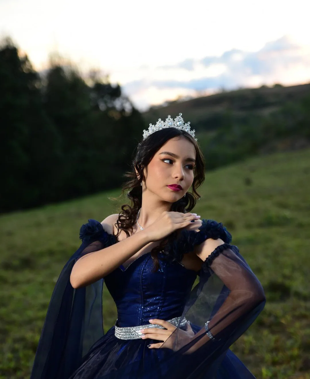 Quinceañera con vestido azul oscuro y tiara plateada en pradera al atardecer